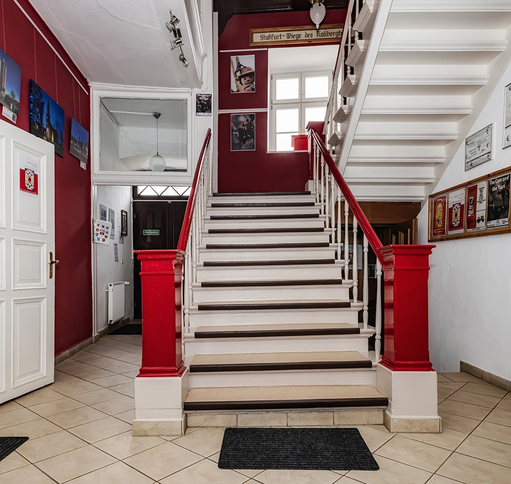 Treppe, mit rotem Geländer und weißen Stufen, beigefarbenen Trittflächen in Eingangshalle, Gemälde und eine Tür zu weiteren Räumen an der linken Wand