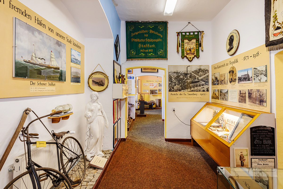 Ausstellungsraum links ein altes Fahrrad und eine weiße Skulptur, rechts an der Wand eine kleine Vitrine, deren Inhalt man nicht sieht